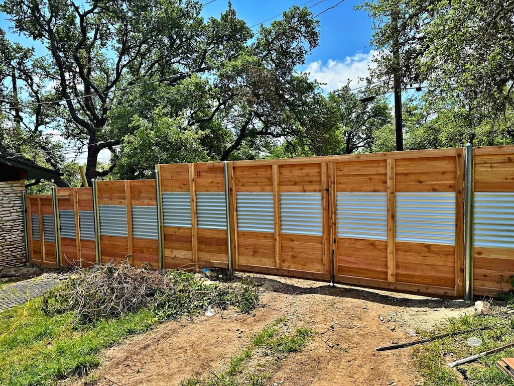 wooden and metal fence divides a grassy yard from lush trees under a blue sky. The fence has corrugated metal panels and wooden frames, creating a rustic yet modern feel.